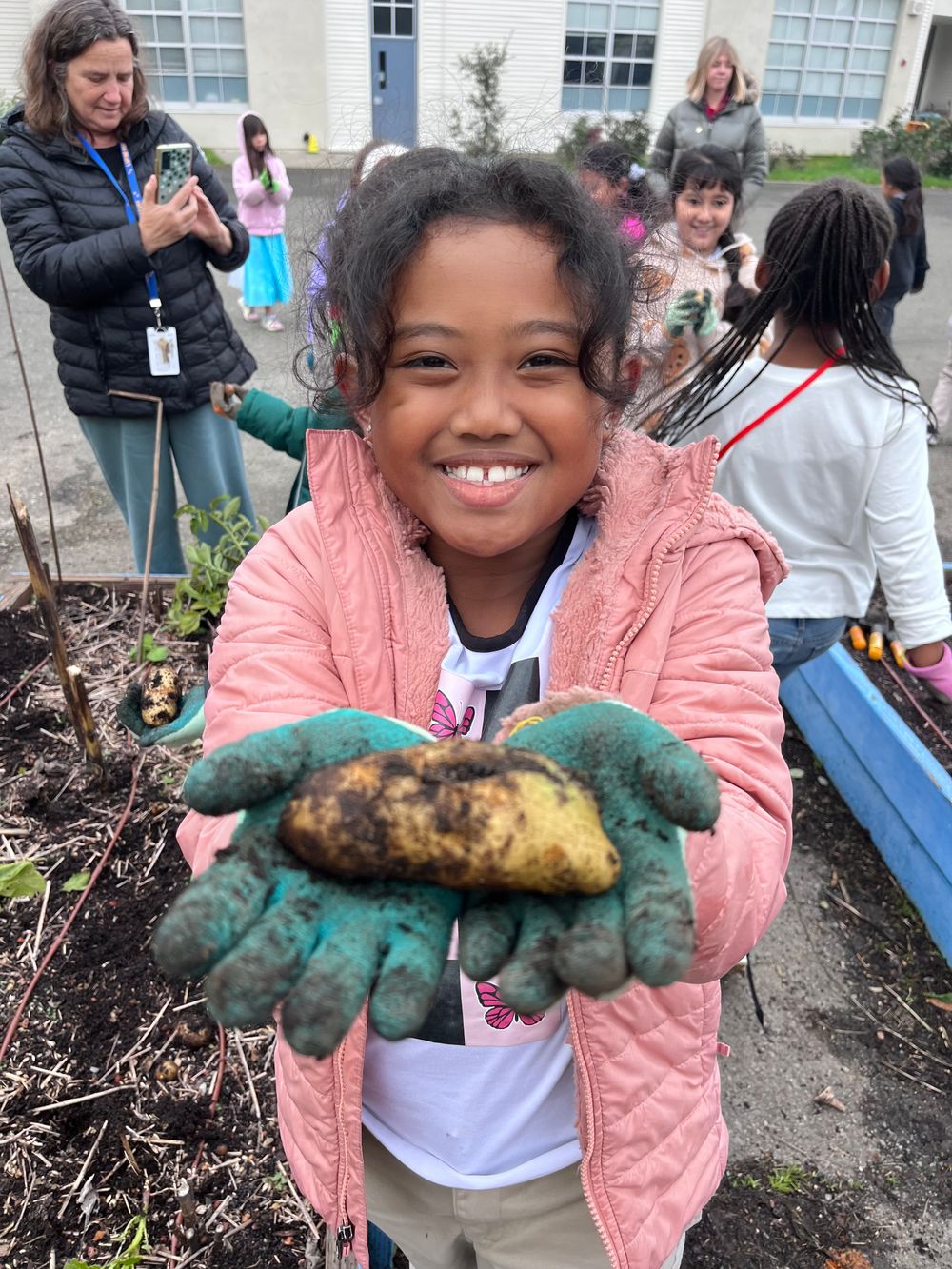 A smiling student in garden gloves holds up a freshly dug root vegetable.