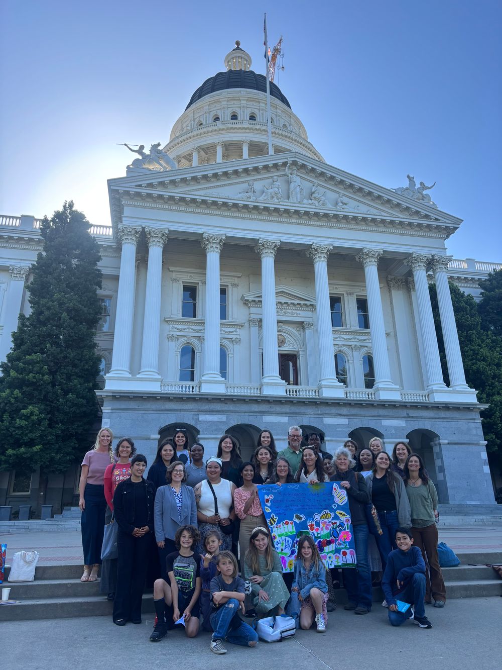 A large group of advocates and children holds a painted banner on the State Capitol steps.