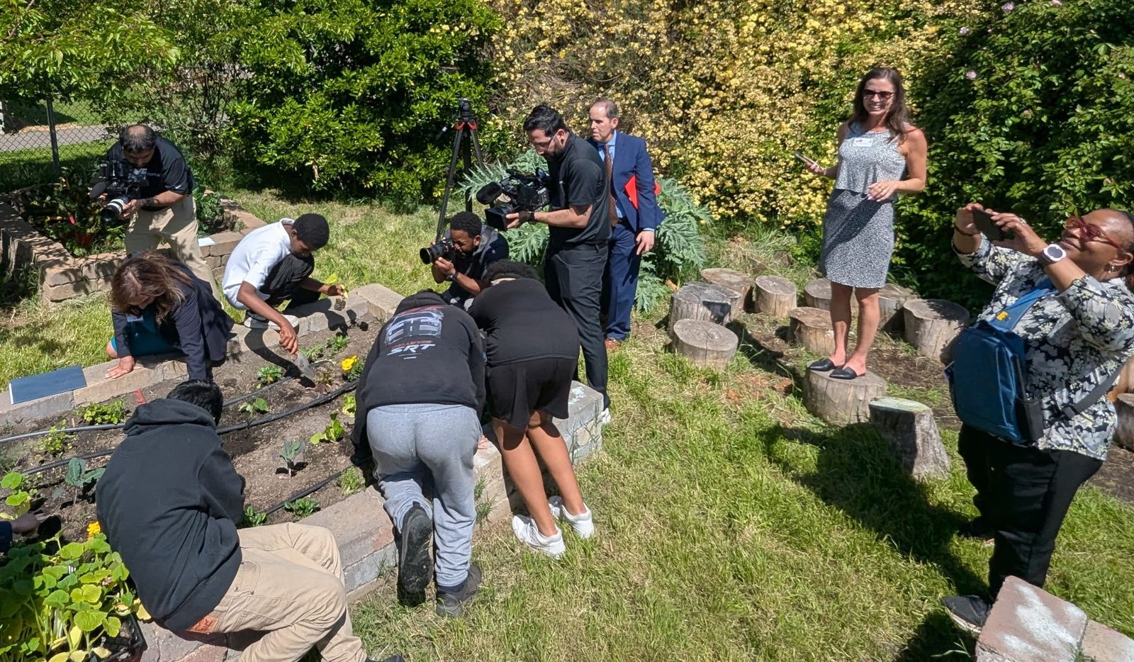 Students plant along a garden bed while camera crews film.